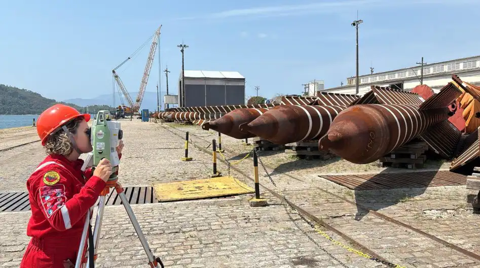 Survey work in a shipyard with a person using equipment and large torpedo anchors lined up on the ground