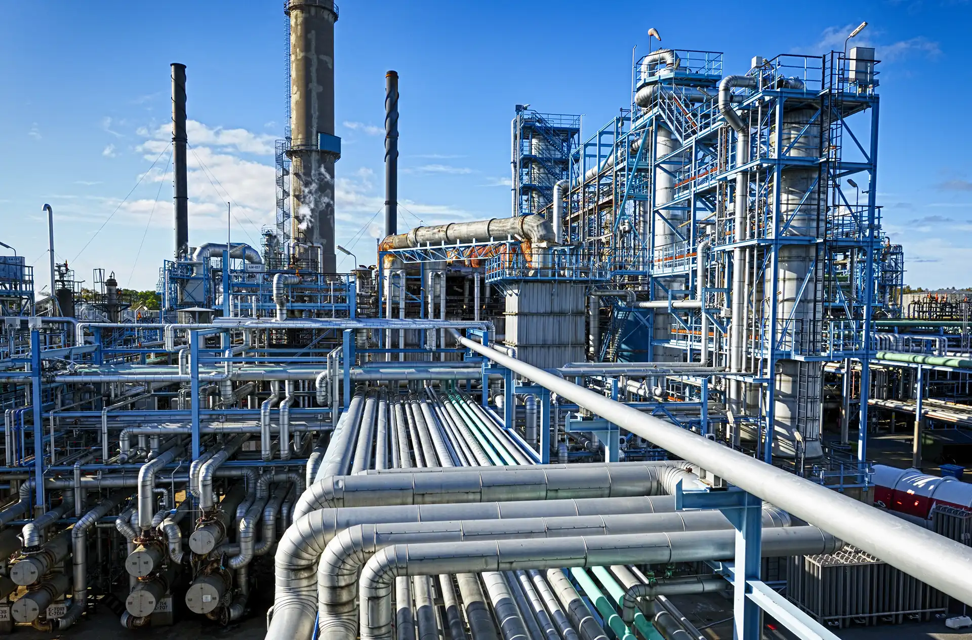 Industrial chemical plant with extensive piping, steel structures, and smokestacks under a clear blue sky.