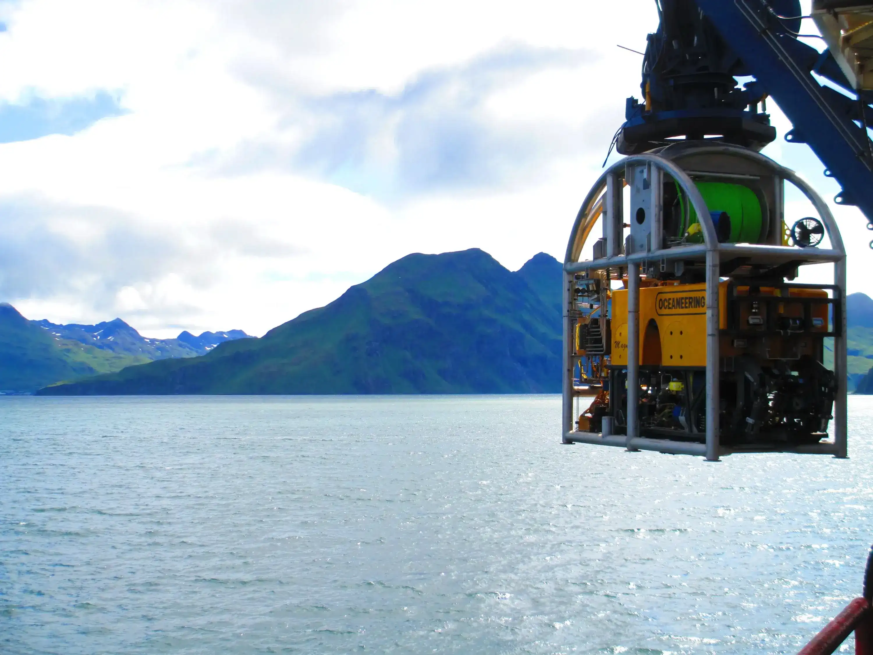 Oceaneering ROV suspended above coastal waters with mountainous shoreline in the background during offshore operations.
