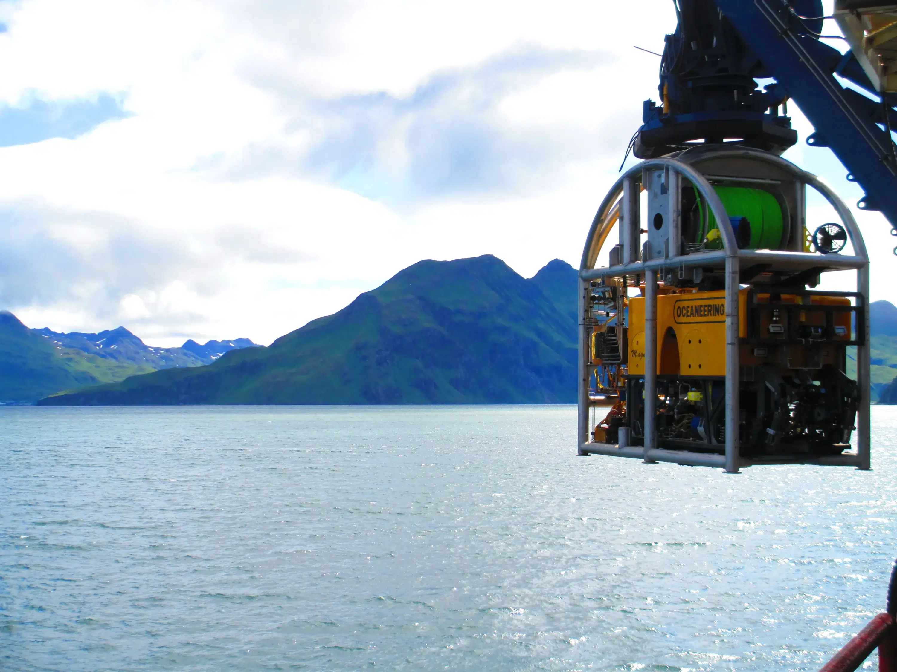 Magnum ROV suspended from a vessel crane above coastal waters with mountains in the background.